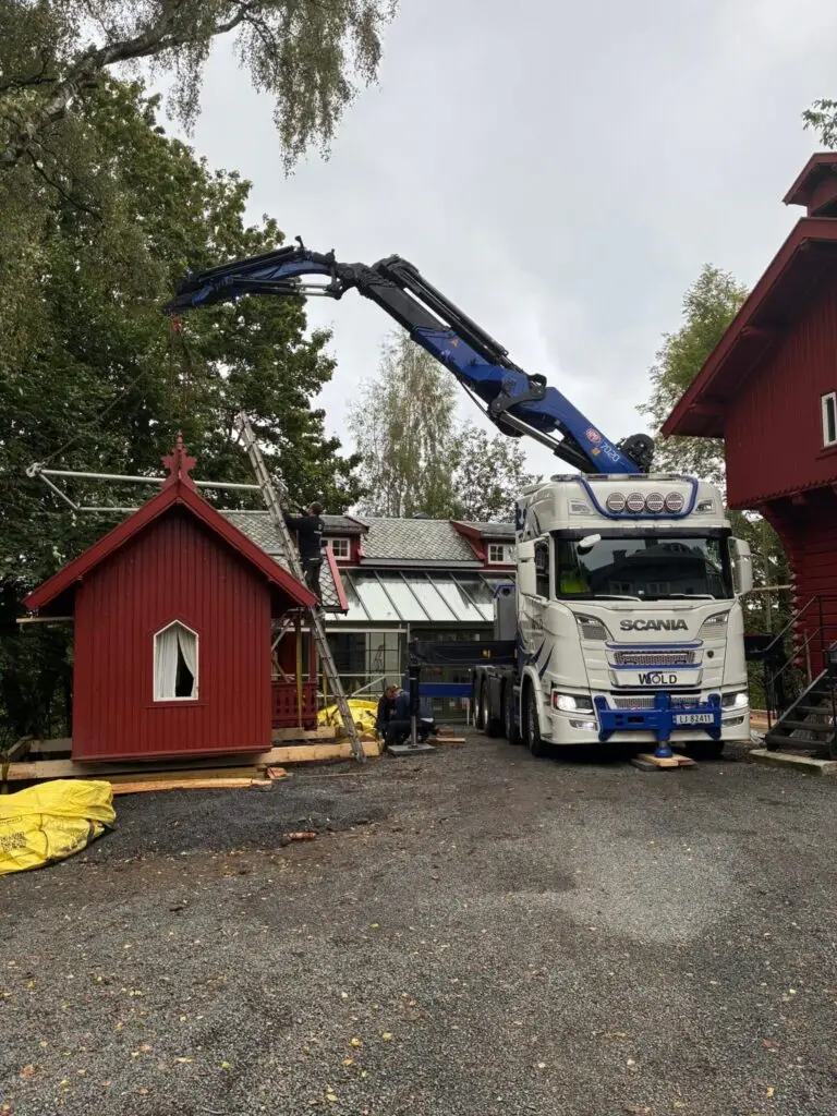 Wold Kran & transports kranbil som løfter et lite rødt hus.